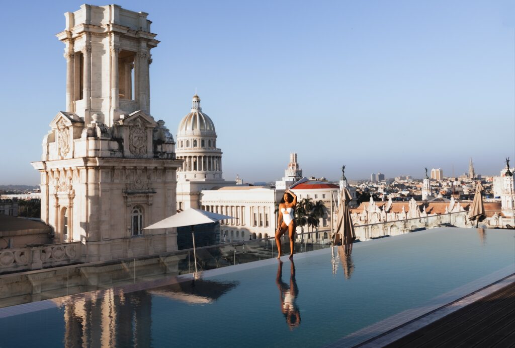 Woman in white swimsuit stands at the edge of an infinity pool overlooking the city in Cuba