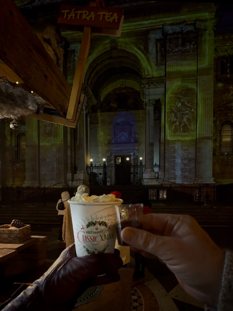 Hands toasting with holiday cheer. Zee holds a hot chocolate and Brett a mulled wine in front of Basilica Church while at the Budapest Christmas Market.
