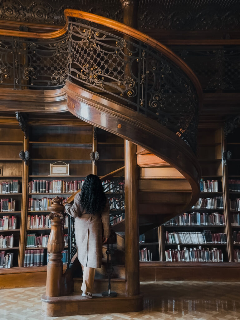 Zee standing beside the ornate spiral staircase at Ervin Szabó Library in Budapest